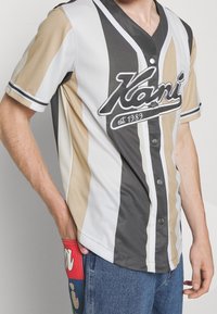Jersey de manga corta a rayas en beige, gris y blanco con escote en V y botones. Logo "Kani est. 1989" en el frente, con acentos azules y rojos en los jeans.