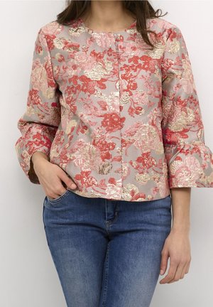 Donna che indossa una giacca in broccato floreale con motivi rossi e oro e jeans blu, con una mano in tasca, in piedi davanti a uno sfondo semplice.