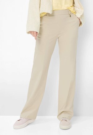 Person iført beige bukser med vide ben, lys gul top, struktureret cremefarvet jakke og lyserøde sneakers, stående mod en hvid baggrund.