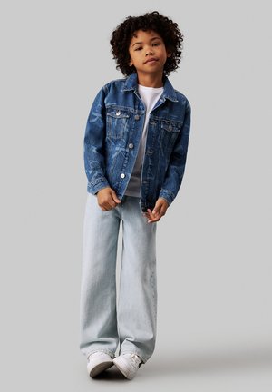 Giacca di denim di un blu medio con un taglio classico, chiusura a bottoni e due tasche sul petto. Abbinata a jeans a gamba larga di un blu chiaro e sneakers bianche.