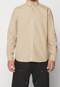 Camisa de botones de manga larga beige con cuello, con botones marrones y un sutil logo en el pecho. Tejido suave con una textura lisa.