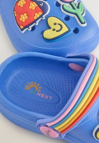 Blaue Gummiclogs mit bunten 3D-Dekorationen: einem Marienkäfer, einem Herz, einem Kaktus und einem Regenbogenriemen. Profiliertes Fußbett mit Markendetails im Inneren.