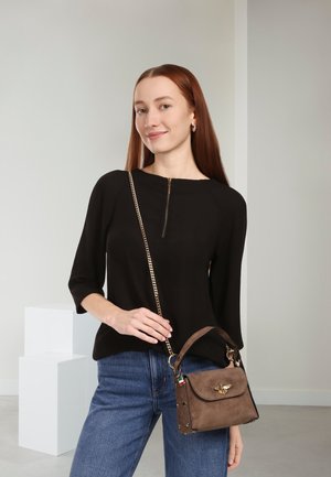 Zwarte blouse met een rits bij de halslijn, gecombineerd met blauwe jeans. Houdt een bruine suède handtas vast met gouden hardware en een bijenmotief als detail.