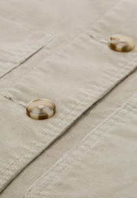 Beige fabric with a textured surface featuring two round, light brown buttons with a subtle pattern and stitching detail.