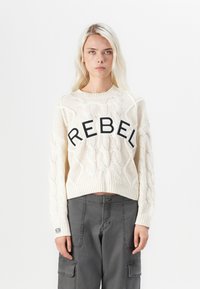 Pulôver curto em malha de creme, apresentando um gráfico preto proeminente "REBEL" na parte da frente. Tecido texturizado com punhos e bainha canelados.