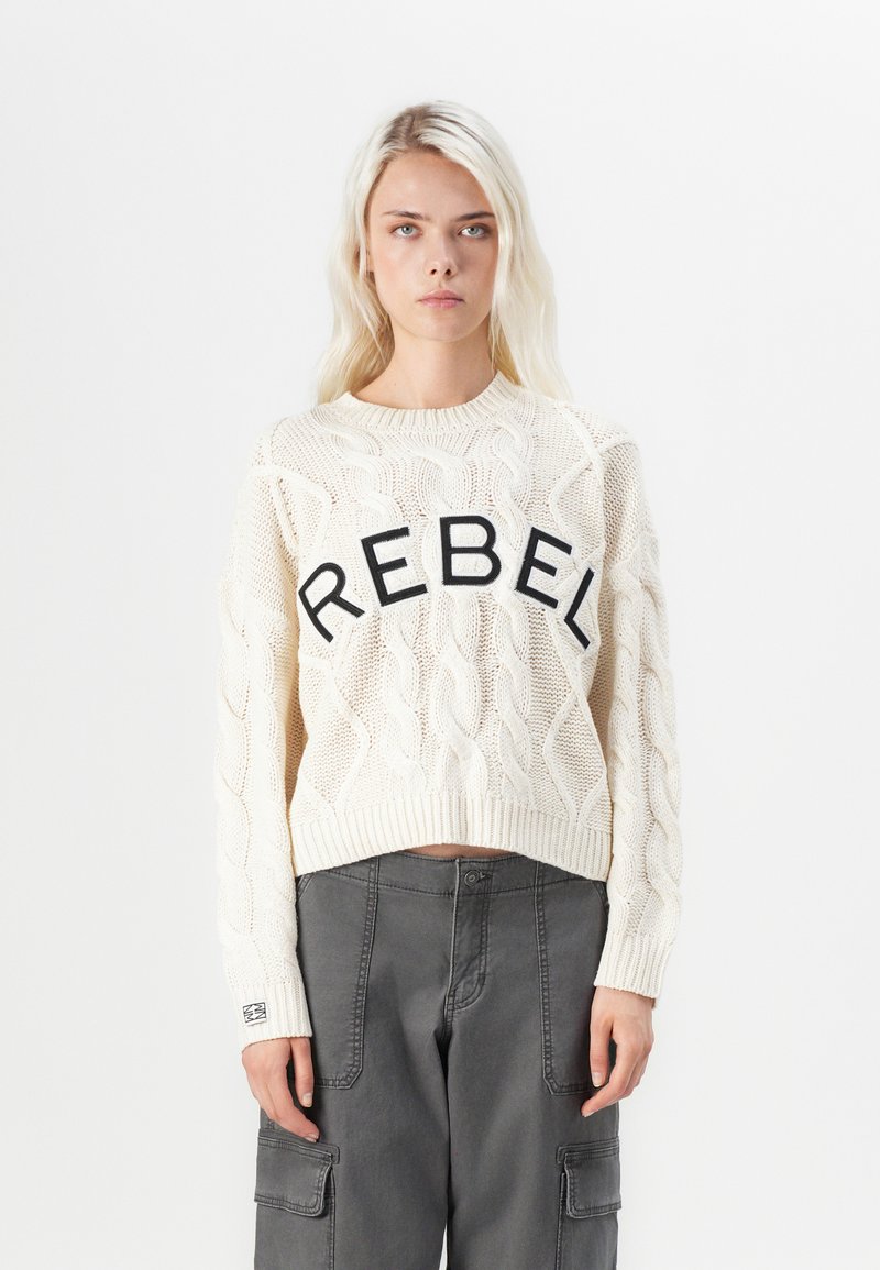 Pulôver curto em malha de creme, apresentando um gráfico preto proeminente "REBEL" na parte da frente. Tecido texturizado com punhos e bainha canelados.