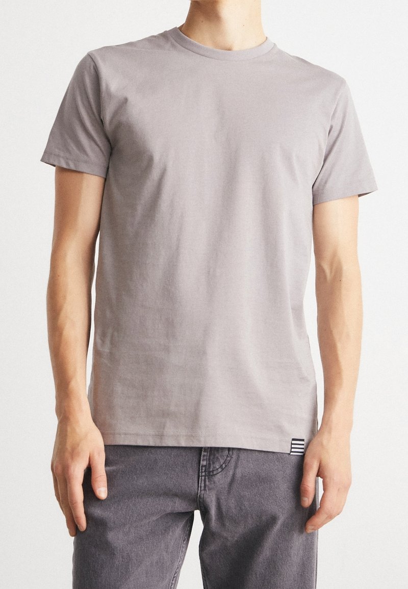 T-shirt en coton gris à manches courtes, encolure ronde, avec un petit accent rayé en bas à gauche de l'ourlet. Texture douce, coupe regular.