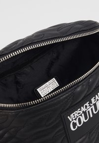 Svart kviltad läderpouch med en silverdragkedja, med en insida märkt "VERSACE JEANS COUTURE" och en slät yta.