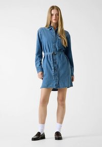 Robe chemise en denim bleu moyen avec manches longues, fermeture boutonnée sur le devant, ceinture à la taille et poche sur la poitrine gauche. Chaussures plates noires portées.