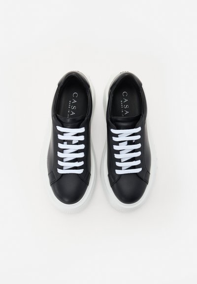 Black leather sneakers with white laces, padded collar, and white rubber sole. Features logo on the insole and smooth texture.