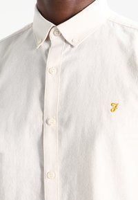 Beige Hemd zum Knöpfen mit einem kleinen gelben, gestickten Logo, einem Kragen und leicht strukturierter Stoff.