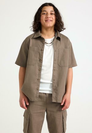 Ung person med skulderlangt krøllet hår iført brun kortærmet skjorte med knapper og cargobukser over en hvid t-shirt, smilende.