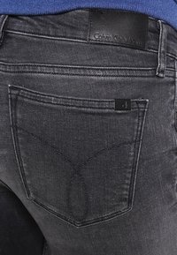 Jeans de mezclilla negros con un diseño de bolsillo trasero que muestra detalles de costura, una etiqueta de marca de cuero negro y una pequeña etiqueta de logo.