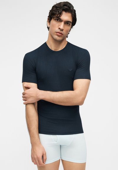 Νέος άνδρας με σκούρα μαλλιά, που φοράει εφαρμοστό σκούρο μπλε κοντομάνικο πουκάμισο και ανοιχτό μπλε μποξεράκια, ποζάρει με σταυρωμένα τα χέρια.