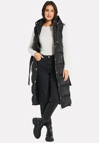 Schwarze lange Pufferweste mit Kapuze, Reißverschluss und Druckknopfverschluss, seitlichen Taschen und goldfarbenen Beschlägen, über einem weißen Oberteil und schwarzen Hosen getragen.