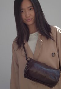 Giovane donna con lunghi capelli scuri, vestita con un cappotto beige e una maglietta bianca, che porta una borsa a tracolla in pelle marrone scuro sul petto.