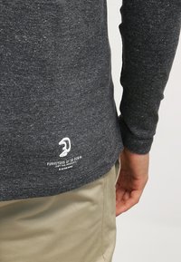 Mörkgrå långärmad skjorta med en fläckig struktur, med vit text/logotyp vid fållen. Materialet verkar vara en bomullsblandning.