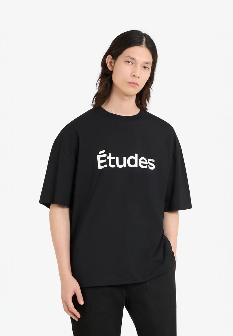 T-shirt noir en coton oversized avec un col rond et des manches courtes. Présente un logo blanc "Études" en évidence sur le devant.