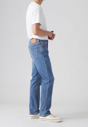 Homme portant un jean bleu, un t-shirt blanc et des baskets blanches, debout de profil avec les mains jointes devant sur un fond uni.