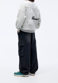 Persona che indossa un felpa grigia chiara con grafiche a forma di stella e testo, pantaloni cargo neri larghi e sneakers turchesi, in piedi con le mani in tasca.