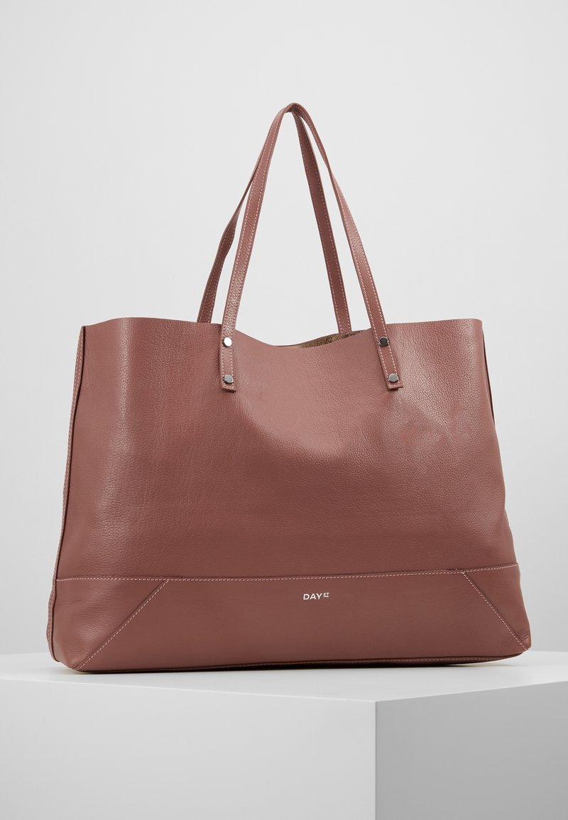 Bruine leren draagtas met een getextureerde afwerking, twee schouderbanden en een logo onderaan. Heeft een ruim interieur en zachte randen.