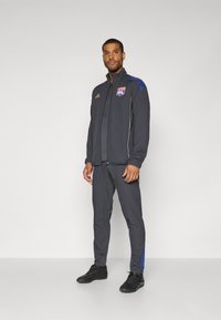 Sport joggedress i mørkegrått med blå detaljer; jakke med glidelås og logo samt gull detaljer, kombinert med tettsittende bukser og svarte treningssko.
