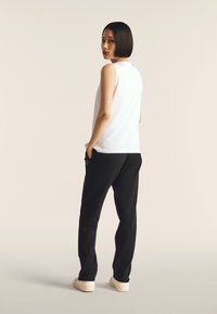 Top bianco senza maniche abbinato a pantaloni della tuta neri. I pantaloni presentano tasche laterali e un logo. Il modello si trova di spalle contro uno sfondo neutro.
