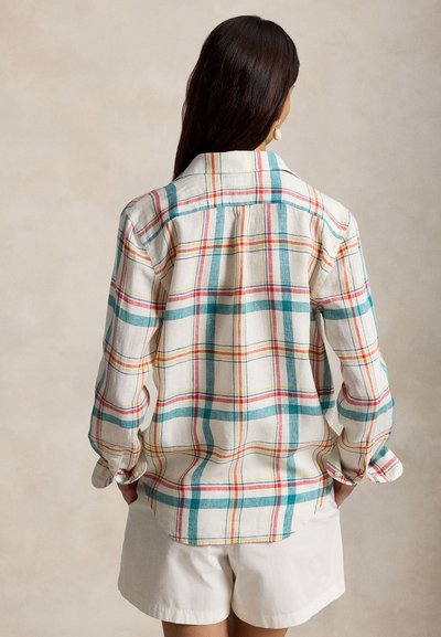 Kvinne med langt mørkt hår iført en beige rutete skjorte med lange ermer, med grønne, røde og gule striper, og hvite shorts, stående med hendene i lommene.