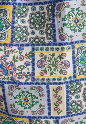 Textilie s patchworkovým vzorem, který kombinuje květinové a geometrické motivy v zelené, modré, žluté a oranžové na světlém pozadí.