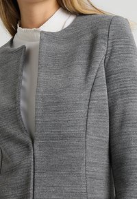 Graue, strukturierte Jacke mit rundem Ausschnitt, minimalistischem Design und glattem Innenstoff. Keine sichtbaren Knöpfe oder Verzierungen.