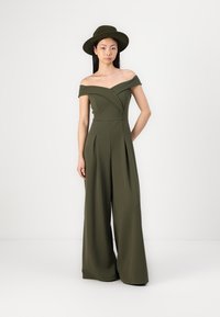 Jumpsuit verde oliva off-the-shoulder in un morbido tessuto; design a gamba larga, con pieghe e un corpetto aderente. Indossato con un cappello abbinato.