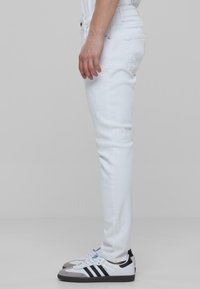 Jeans de denim blanco con un ajuste slim, que presentan un ligero desgaste, diseño de cinco bolsillos y cierre de botón estándar, combinados con zapatillas grises y blancas.