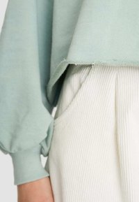 Sudadera verde menta con dobladillo sin terminar y mangas abullonadas, superpuesta sobre unos pantalones acanalados blancos con bolsillos laterales y detalles plisados.