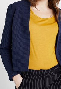 Laivastonsininen teksturoitu bleiseri, keltainen scoop-kauluksellinen toppi ja mustat raidalliset housut. Bleiserissä on jämäkkä muotoilu, jossa on uritettu pinta.