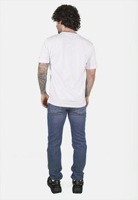 T-shirt bianca in cotone con maniche corte, abbinata a jeans in denim blu. Il modello è in piedi su uno sfondo neutro, mostrando la vestibilità dell'outfit.