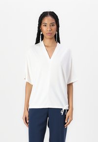 Witte top met korte mouwen, een V-hals en een trekkoorddetail aan de onderkant. Soepele textuur en een relaxte pasvorm. Gecombineerd met donkerblauwe broek.