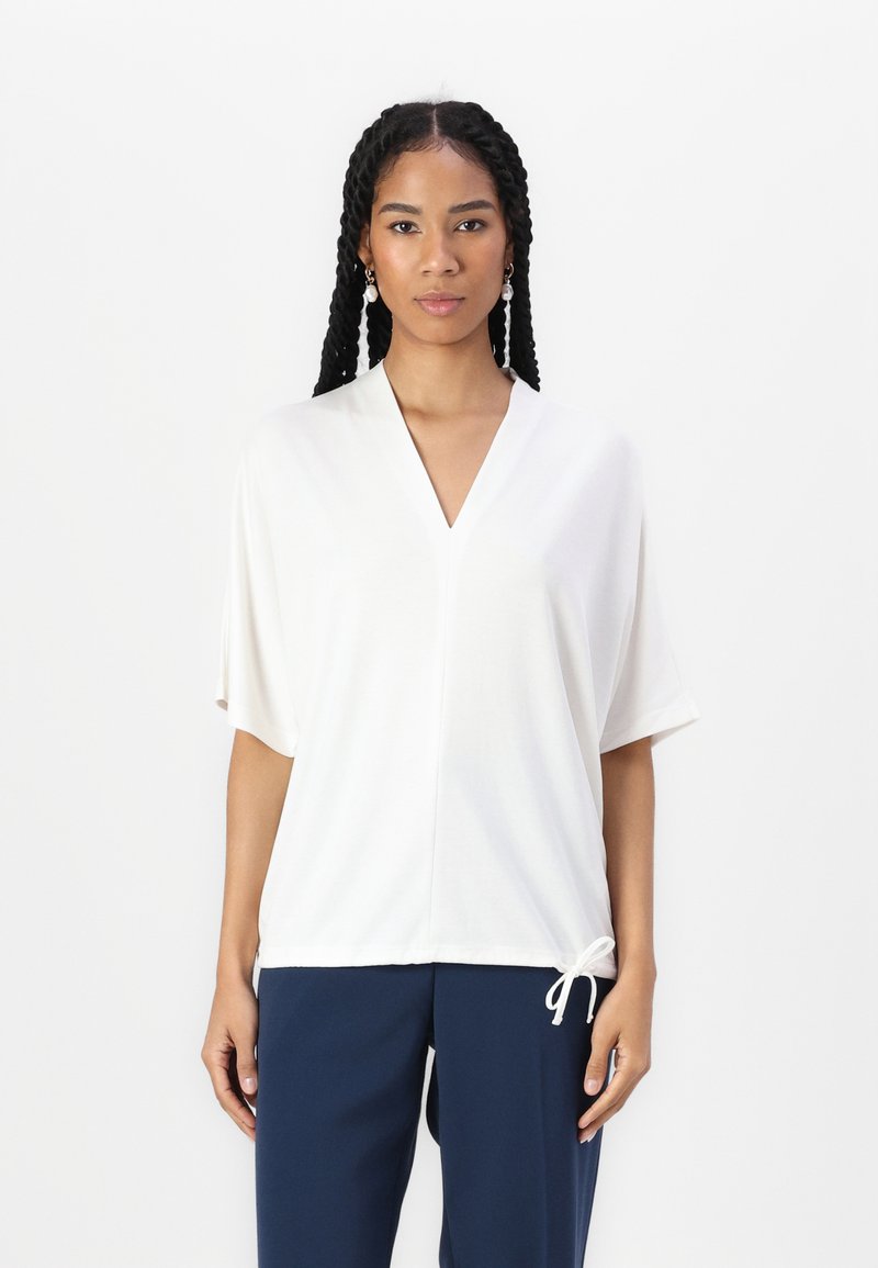 Witte top met korte mouwen, een V-hals en een trekkoorddetail aan de onderkant. Soepele textuur en een relaxte pasvorm. Gecombineerd met donkerblauwe broek.