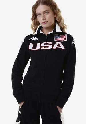 Giovane donna con giacca nera con zip, con la scritta "USA" e una toppa con la bandiera americana, le mani nelle tasche, espressione neutra.