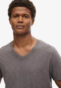 Camiseta gris de cuello en V, fabricada con suave algodón. Presenta mangas cortas y un corte relajado. Textura lisa con un sutil patrón jaspeado.