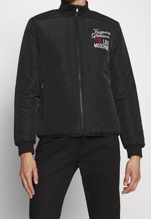 Personne portant une veste noire à fermeture éclair avec "Toujours Glamour Love Moschino" brodé en blanc et rouge sur la poitrine gauche.