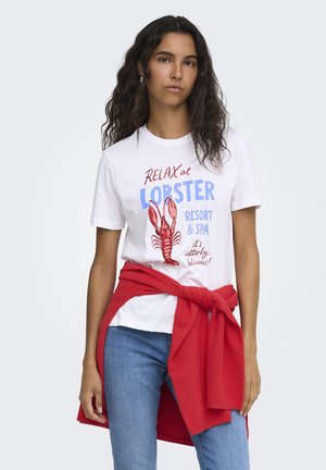 Mujer con cabello largo y rizado que lleva una camiseta blanca con una imagen y texto de una langosta roja, vaqueros azules y un suéter rojo atado alrededor de su cintura.