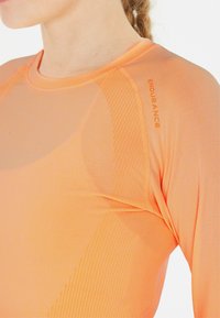 Pitkähihainen urheilupaita kirkkaan oranssina, valmistettu joustavasta kankaasta, jossa on ribbaitua yksityiskohtaa hartioissa ja hienovaraista brändäystä hihassa.