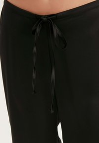 Zwarte loungebroek met een gladde textuur, voorzien van een tailleband met trekkoord dat is vastgeknoopt met satijnen linten. Geen opvallende patronen of accenten zichtbaar.