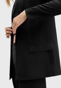 Personne boutonnant un bouton d'un blazer noir sur mesure avec poche à rabat, portant un pantalon noir assorti, sur un fond clair uni.