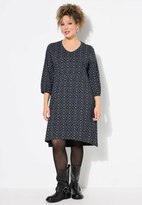 Schwarz-weiß gemustertes Kleid mit dreiviertel Ärmeln, einem V-Ausschnitt und einem weit ausgestellten Saum, kombiniert mit knielangen schwarzen Stiefeln.