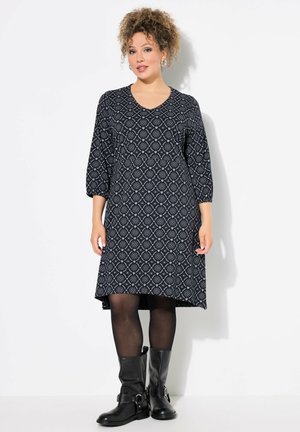 Schwarz-weiß gemustertes Kleid mit dreiviertel Ärmeln, einem V-Ausschnitt und einem weit ausgestellten Saum, kombiniert mit knielangen schwarzen Stiefeln.
