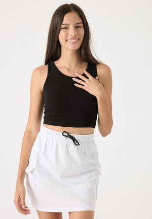 Joven sonriente, lleva un crop top negro sin mangas y una falda blanca con cordón, de pie frente a un fondo claro y simple.