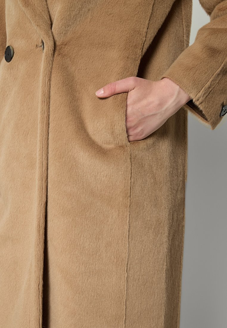 Jasnobrązowy płaszcz uszyty z miękkiej, strukturalnej tkaniny, z guzikowym zapięciem z przodu oraz boczną kieszenią, w której znajduje się ręka.