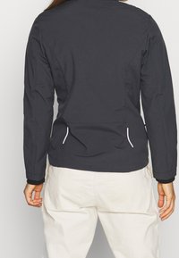 Veste noire à manches longues, dotée de poches latérales, d'une texture lisse et d'accents blancs contrastants le long des poches et des poignets.