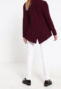 Bordeaux gebreide trui met een hoge kraag en een split aan de onderkant, gecombineerd met witte slim-fit broeken en zwarte instappers.